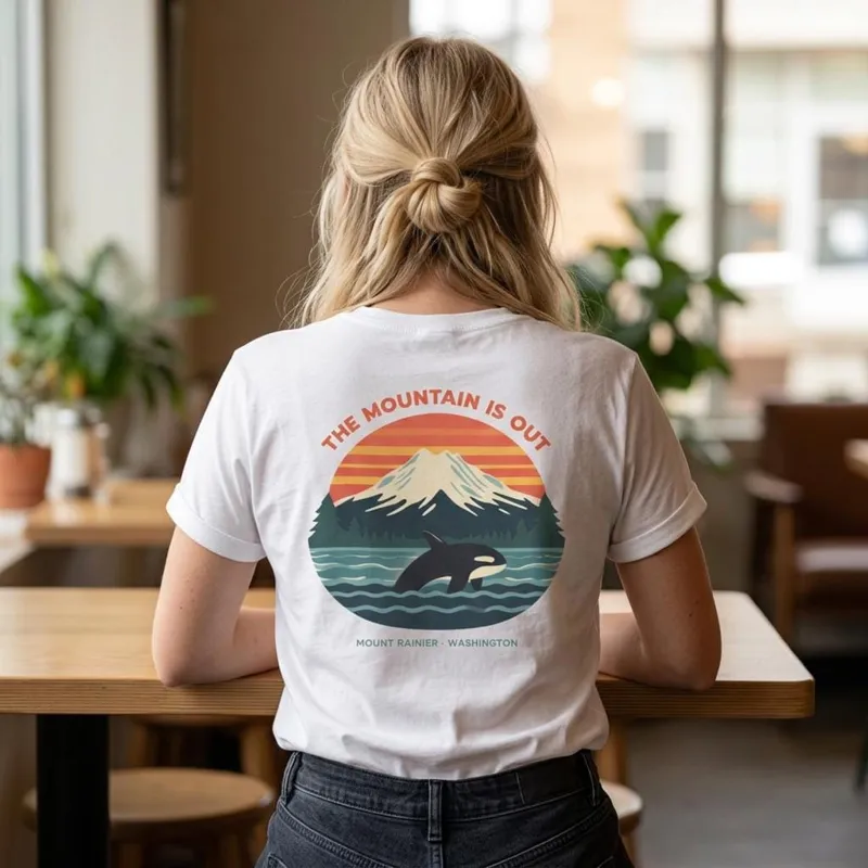 Back of a white t-shirt with a retro sunset, Mount Rainier and orca graphic reading The Mountain Is Out